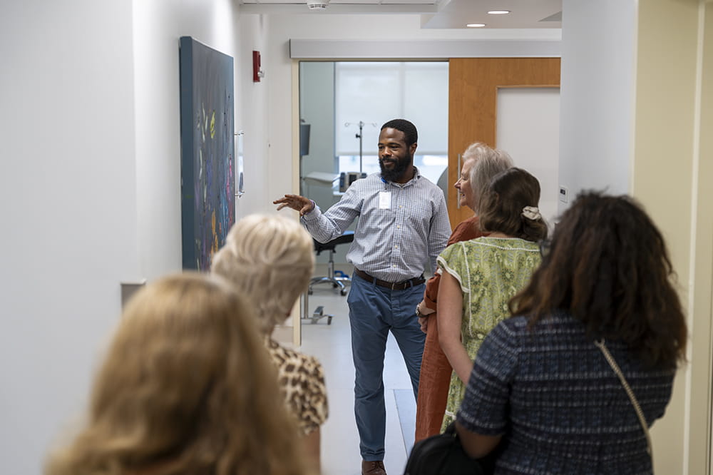 in a clinic hallway, a man in business casual gestures to a painting on the wall while a group of women listen attentively and look at the painting