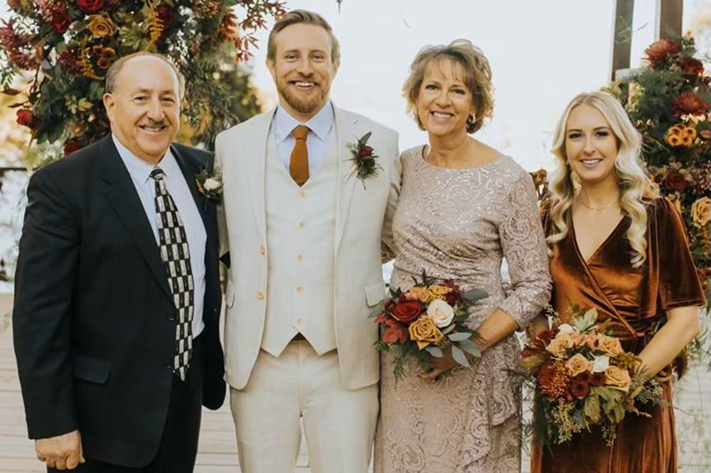 a family wedding photo featuring the father of the groom, the groom in a cream suit with brown tie, the mother of the groom in taupe holding flowers and the sister of the groom in bronze holding flowers