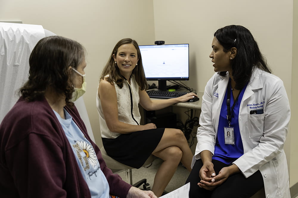 a doctor in a white coat and a patient, sitting facing each other, have their bodies turned to look at a woman sitting at a computer station in the corner of the room who is speaking