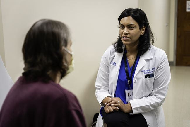 A doctor in a white coat sits to chat with a patient, who has her back facing the camera