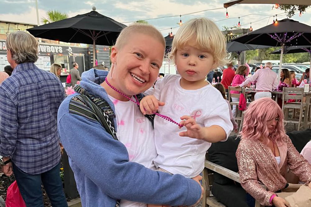 a nearly bald young woman holds a small blond boy, who wears a shirt with a pink ribbon and clasps the pink beads around the woman's neck, at an outdoor event