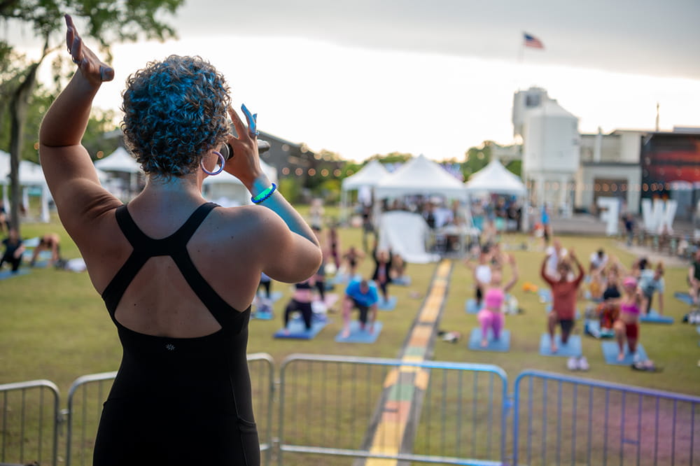 image of the back of a young woman as she speaks into a microphone and gestures with an arm while facing a group of people doing yoga on a large grassy area below