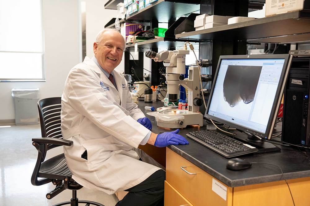 a scientist in a white lab coat sits at the bench next to a microscope with a computer screen showing the image