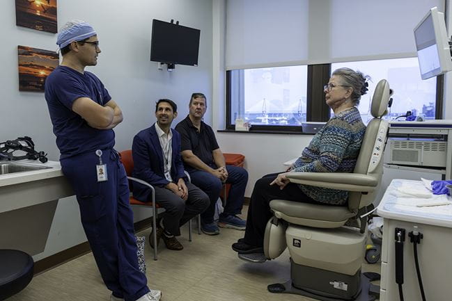 two men in regular chairs and a woman in an exam chair all look to a doctor in dark blue scrubs who leans back against a counter and is talking