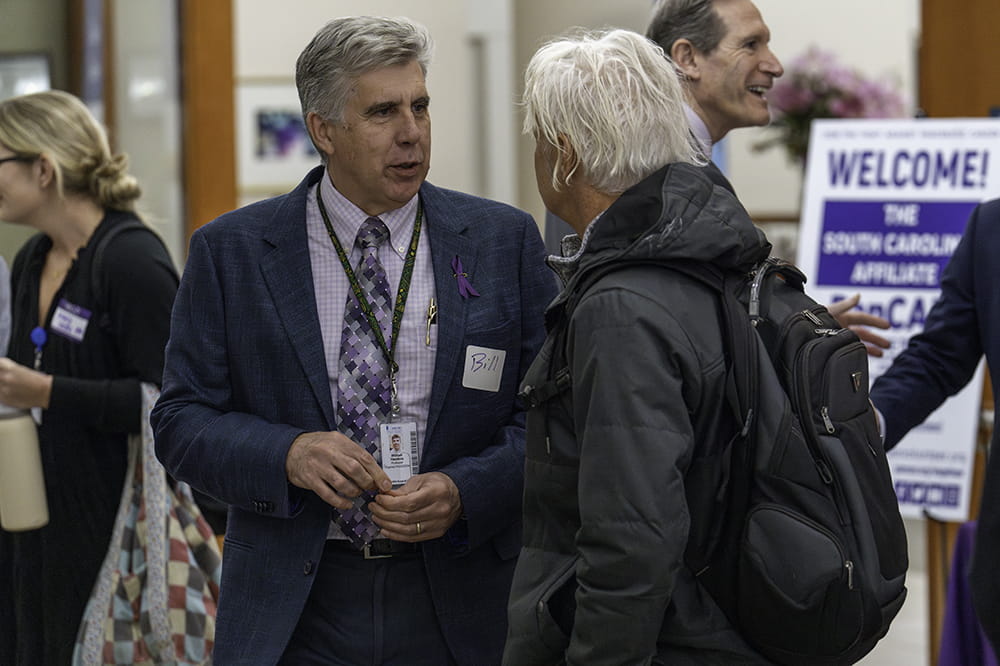 a man wearing purple shirt and tie with a purple ribbon pinned to his lapel talks to another man during a reception at Hollings Cancer Center