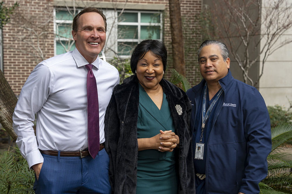 an elegantly dressed woman smiles while standing between a doctor dressed in a tie and button down and a doctor in his scrubs