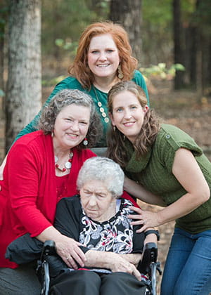  two adult women and their mother lean close to pose with her mother in a wheelchair