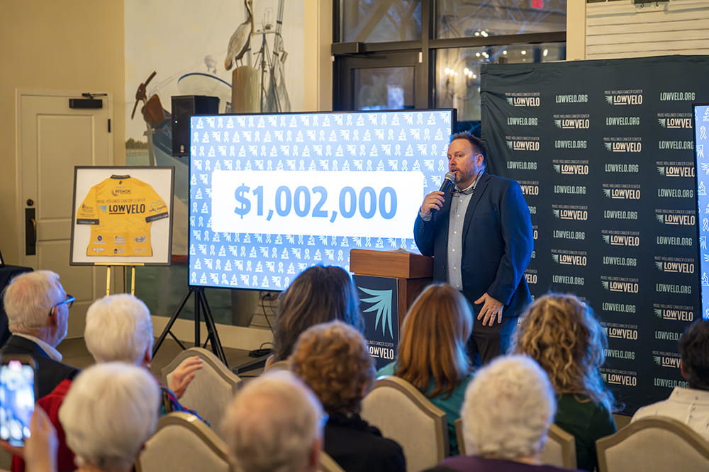 a man stands in front of a crowd with a microphone while a screen behind him displays the dollar figure $1,002,000