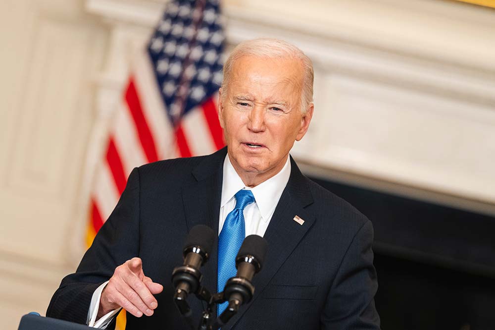 Man in dark suit with blue tie speaks before microphones. An American flag stands behind him.