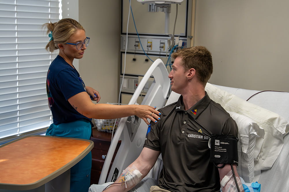 a nurse turns back and lightly touches a young man in a hospital bed on the shoulder as they talk 