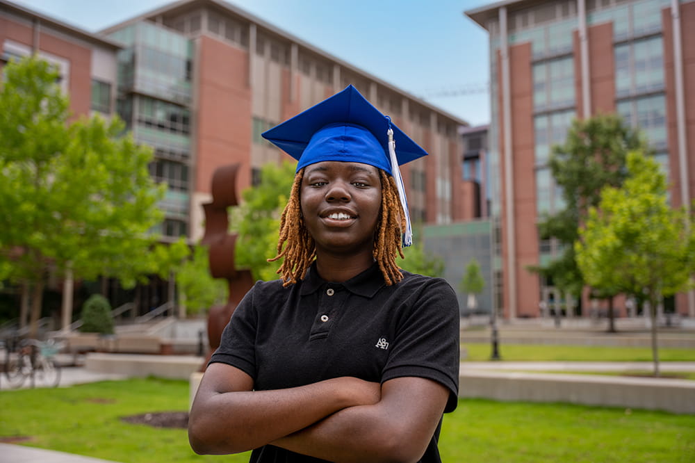 a young woman in a black polo and a blue mortarboard graduation cap poses in front of medical research buildings at MUSC