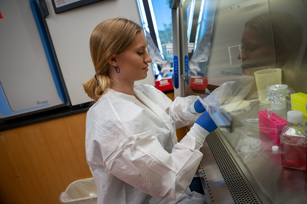 a young woman covered in lab coat and gloves works under a hood in a science lab