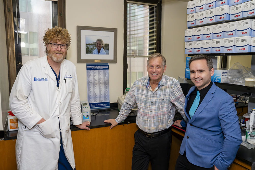 a doctor, donor and researcher pose in a cancer research lab with the photo of the donor's deceased wife on the wall 
