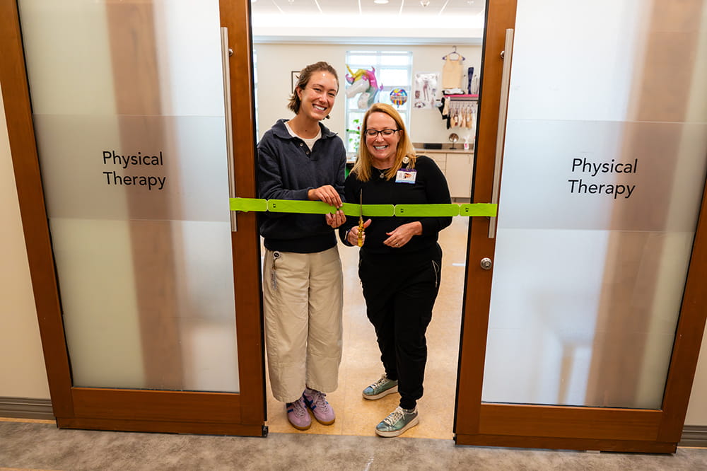two women stand in a doorway prepared to cut a ceremonial ribbon made of physical therapy tape