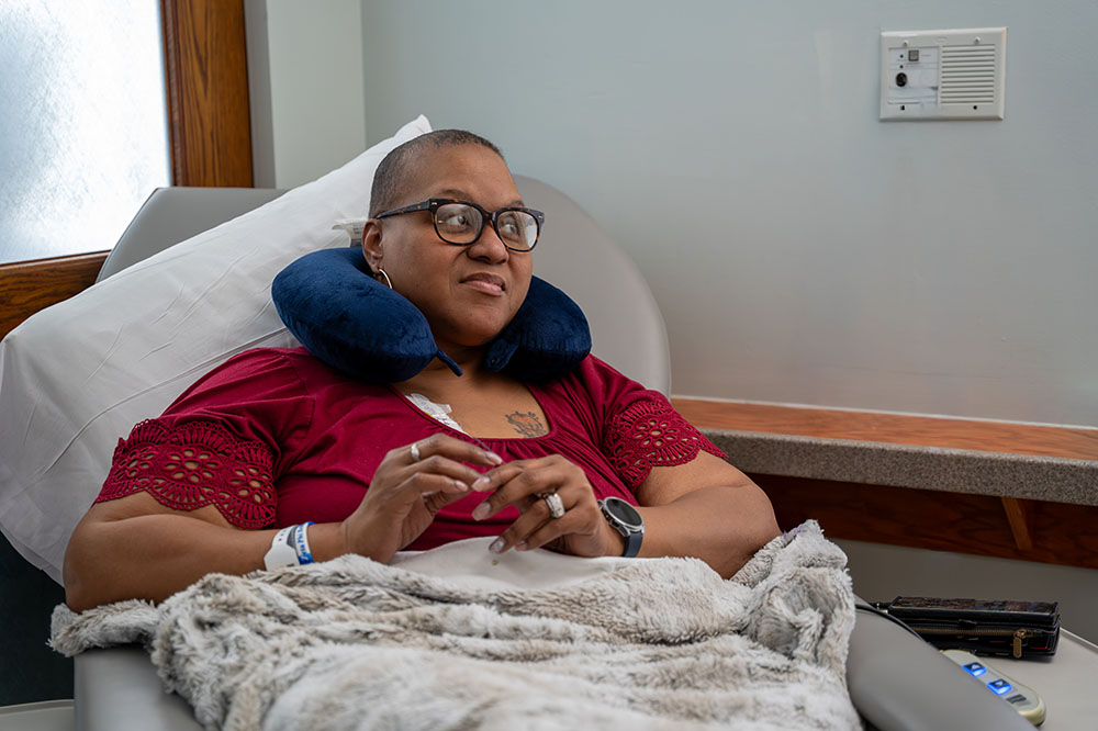 a woman sits in an infusion chair with a neck pillow and regular pillow behind her and blanket over her lap