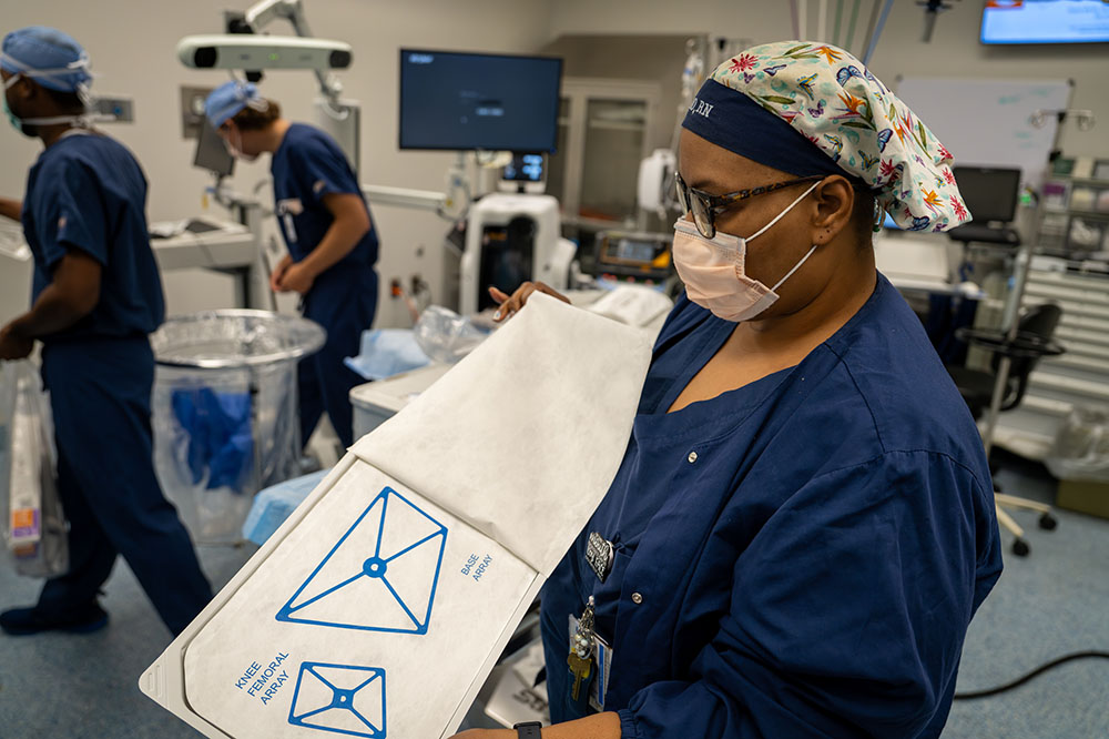 a woman opens a sealed package of operating room materials while two people in the background work on setting up other things