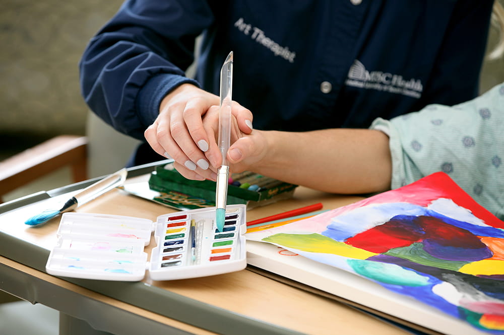 closeup image of a hand guiding another person's hand with a paintbrush on paper